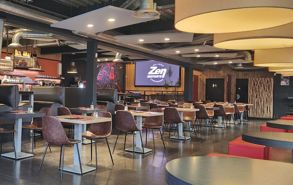 Intérieur du restaurant : tables et chaises, éclairage tamisé, coin bar, écran de projection.