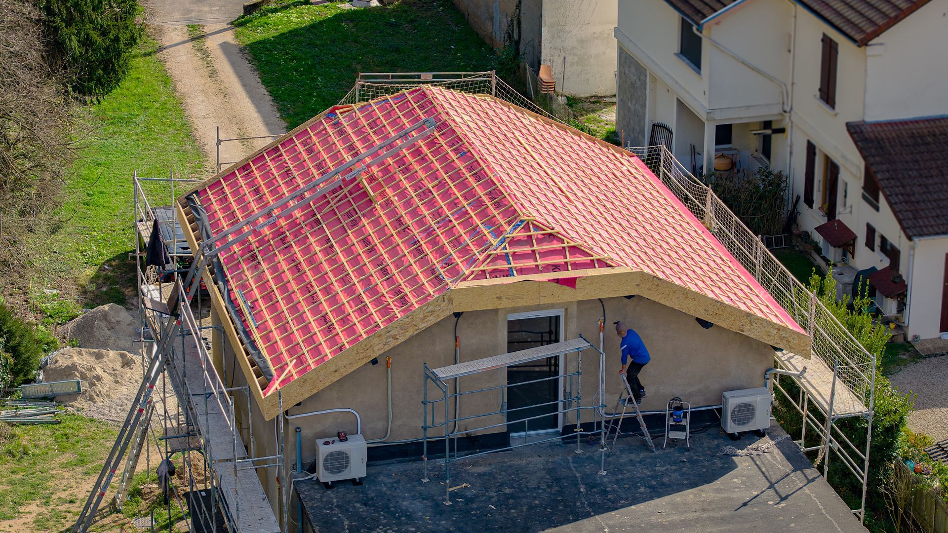 Une petite maison beige au toit de tuiles rouges, vue du ciel, avec une personne debout près du porche.