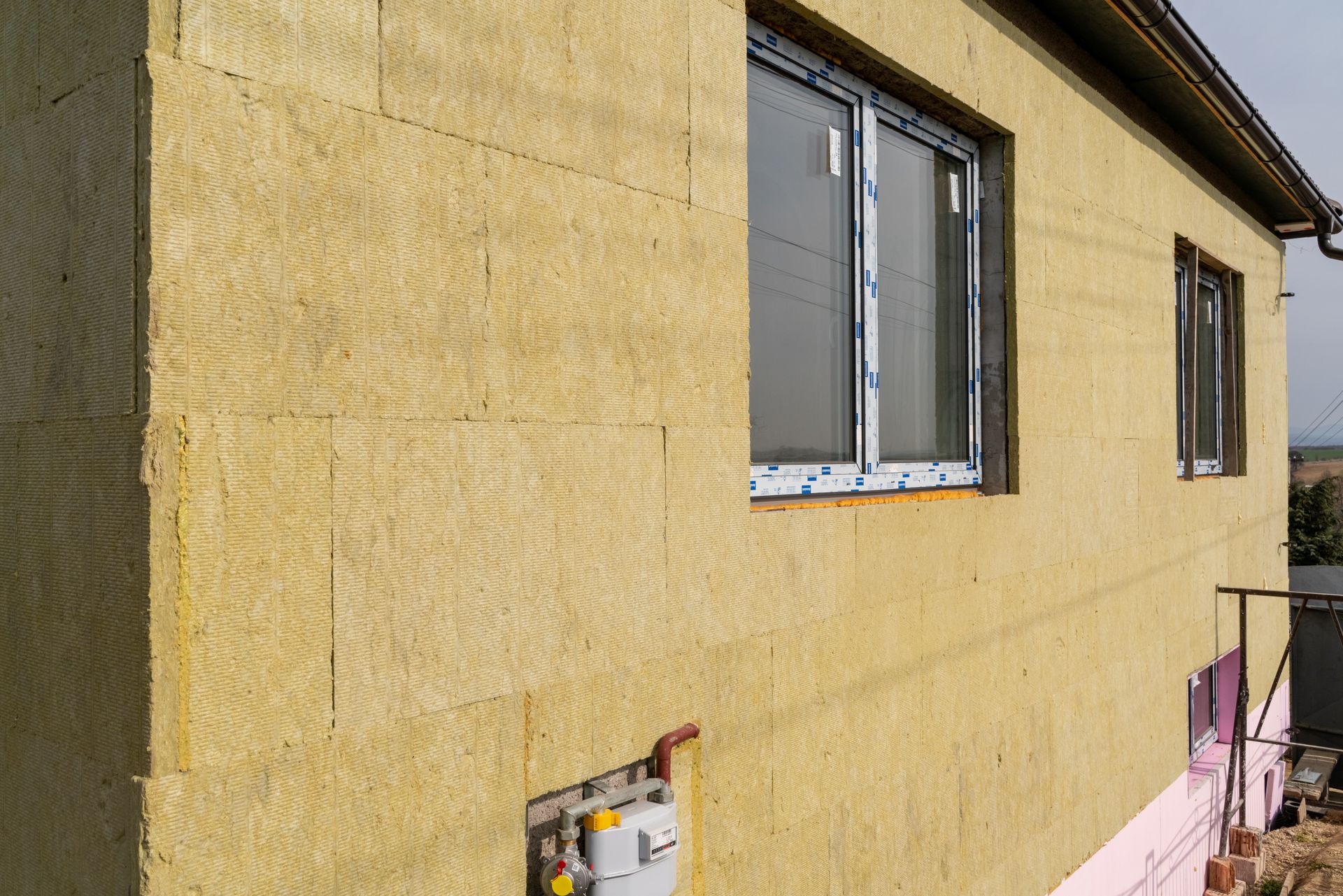 Mur extérieur en stuc jaune avec deux fenêtres et un petit boîtier technique sous la fenêtre de gauche