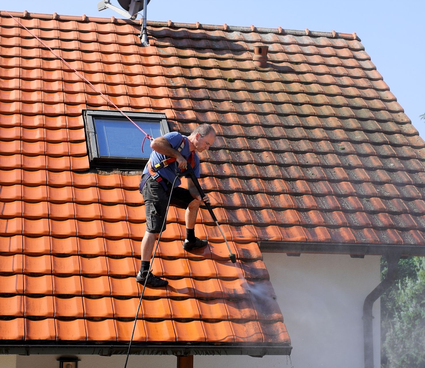 Un ouvrier nettoie au nettoyeur à haute pression un toit de tuiles rouges à côté d'une lucarne et d'une cheminée.