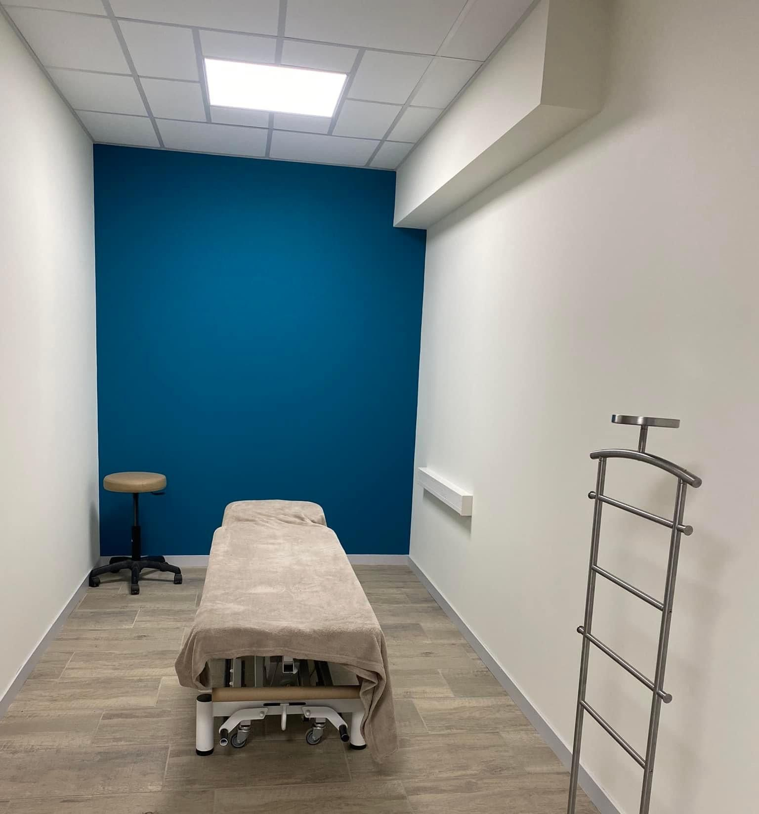 Salle d'examen : mur d'accent bleu, table de massage avec revêtement beige, tabouret, portemanteau, murs et sol neutres.
