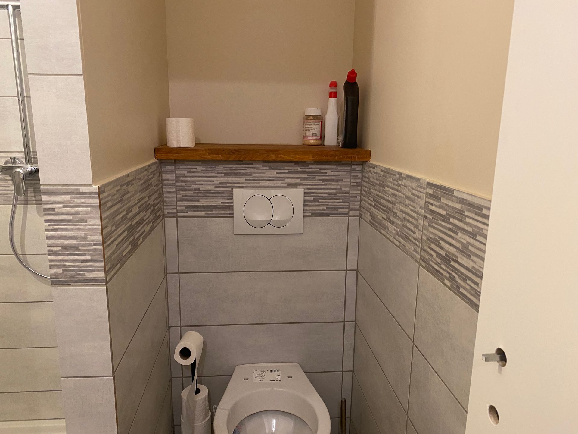 Petite salle de bain avec toilettes, étagère pour produits de nettoyage et carrelage décoratif.