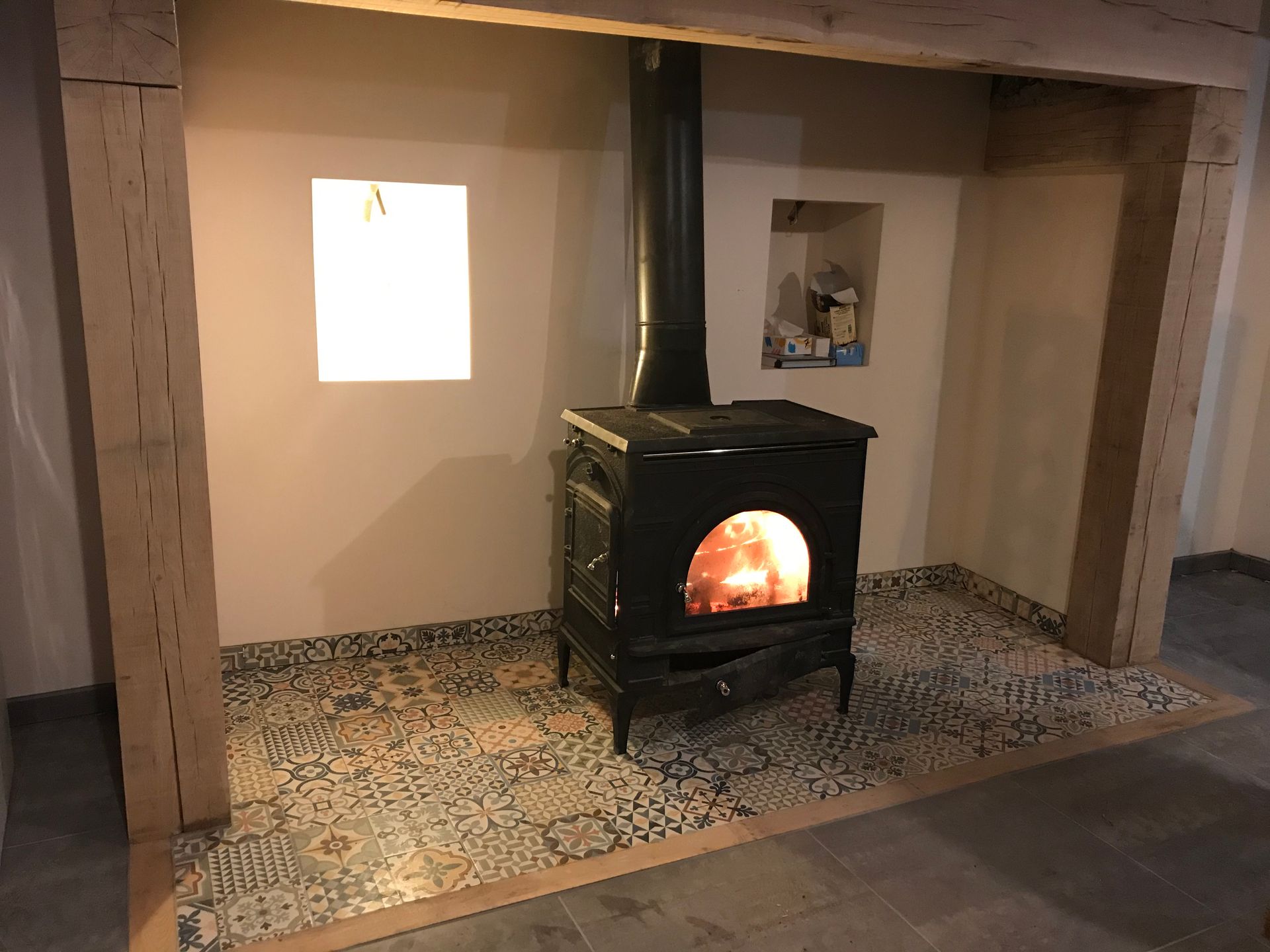 Poêle à bois noir avec feu, installé dans une alcôve avec poutres en bois, sol carrelé et niche encastrée.
