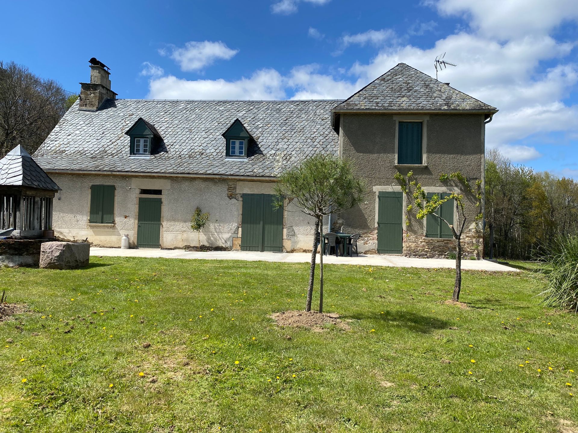 Maison de campagne française aux volets verts, située dans une cour herbeuse, journée ensoleillée.