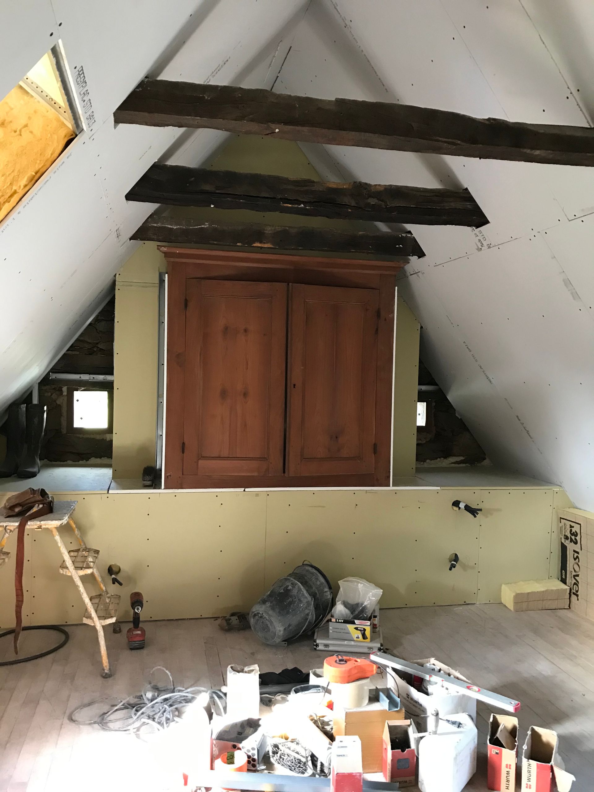 Armoire en bois aménagée dans un grenier, sous poutres apparentes ; matériaux de construction éparpillés au sol.