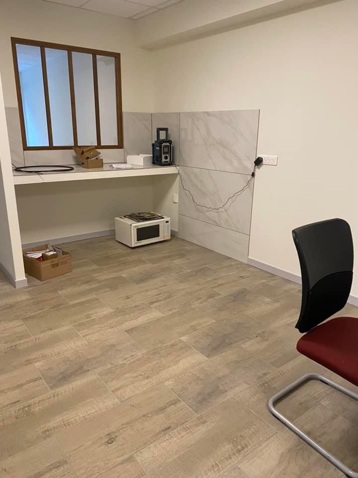 Pièce vide avec fenêtre, comptoir et mur carrelé. Un micro-ondes et d'autres appareils sont présents. Une chaise trône au premier plan.