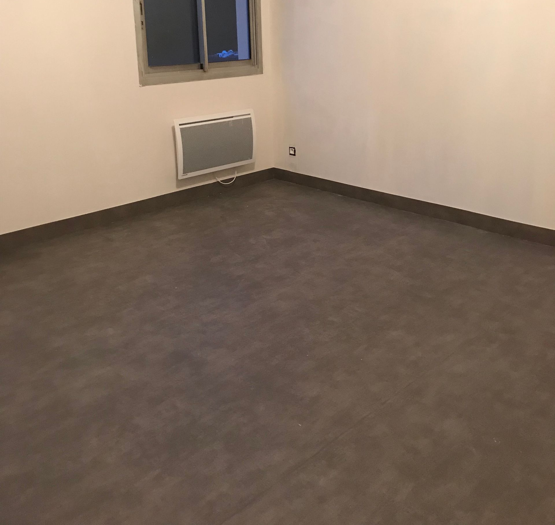 Pièce vide avec sol gris, murs beiges, fenêtre et radiateur mural.
