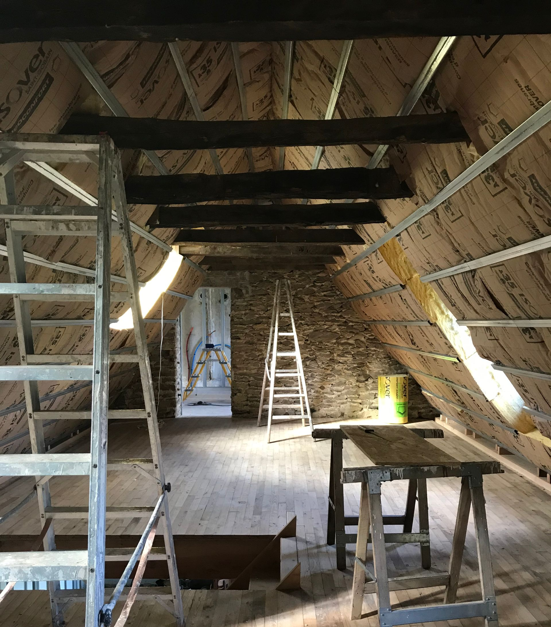 Rénovation de combles en cours : poutres en bois, isolation, échelles et une porte dans un bâtiment rustique.