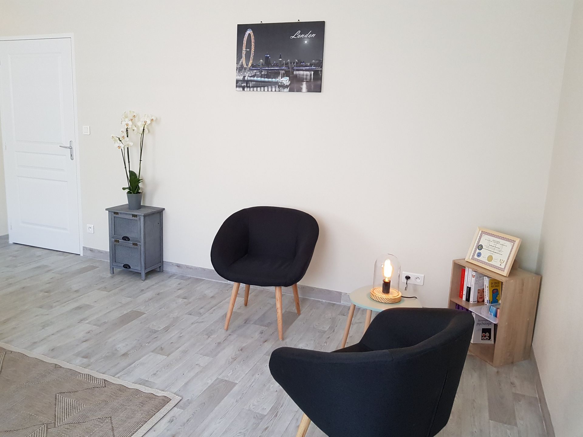 Deux fauteuils noirs dans une pièce bien éclairée, table, petite bibliothèque, avec une photo de paysage urbain au mur.