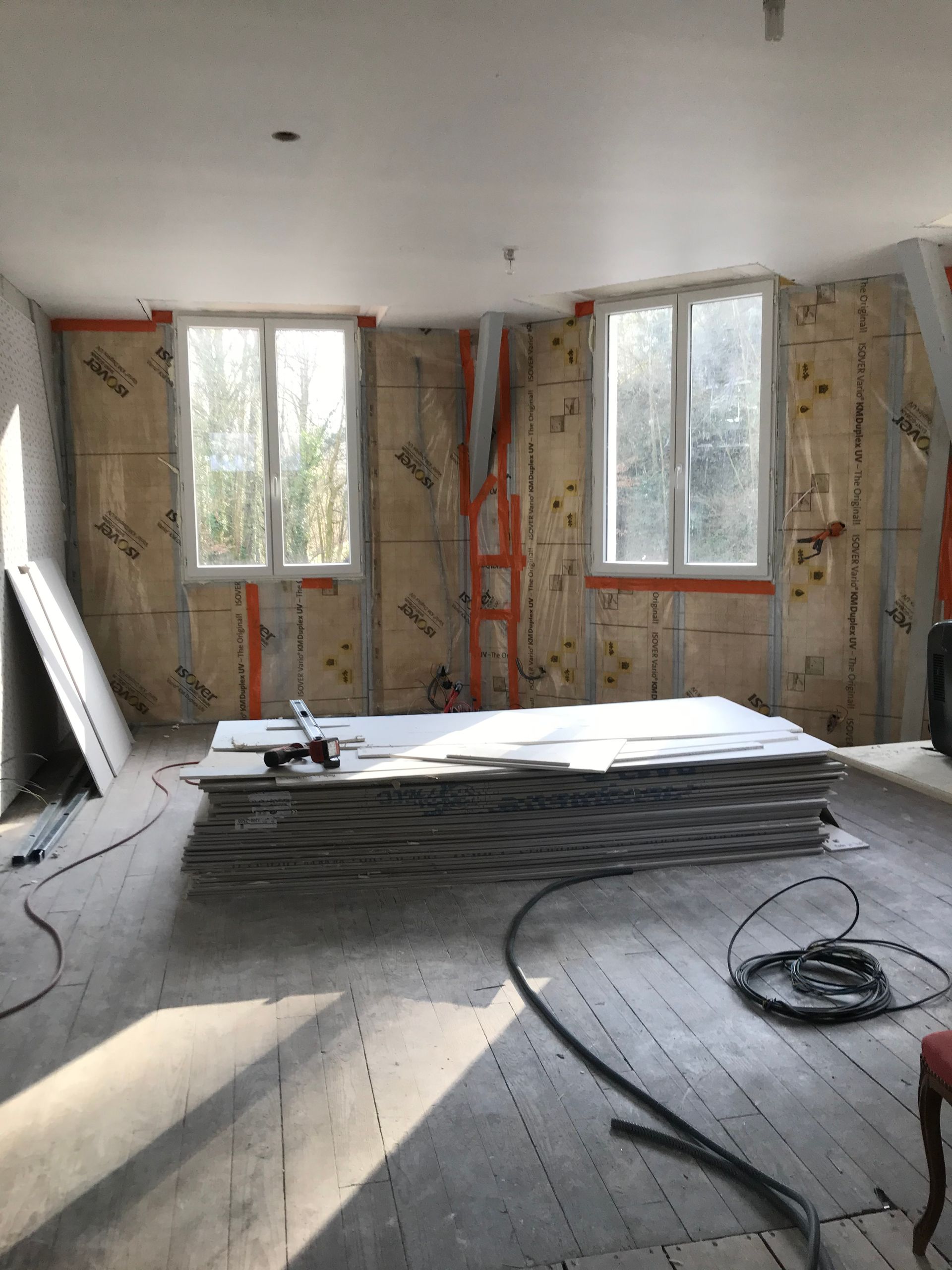 Intérieur d'une pièce en construction, avec isolation apparente, fenêtres et plaques de plâtre empilées sur le sol.