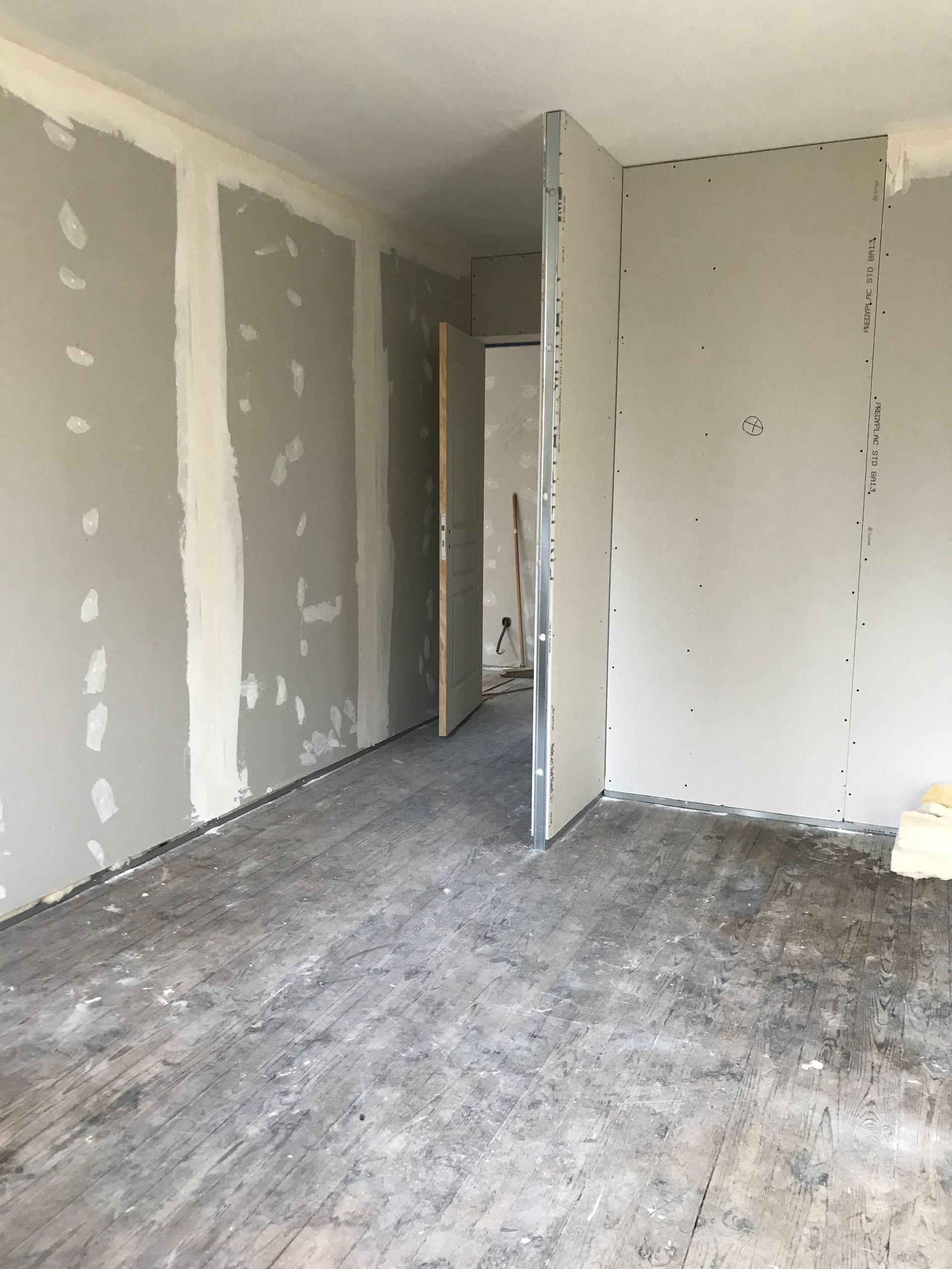 Chambre en construction : cloisons sèches installées, murs gris, porte menant à une autre pièce, plancher non fini.