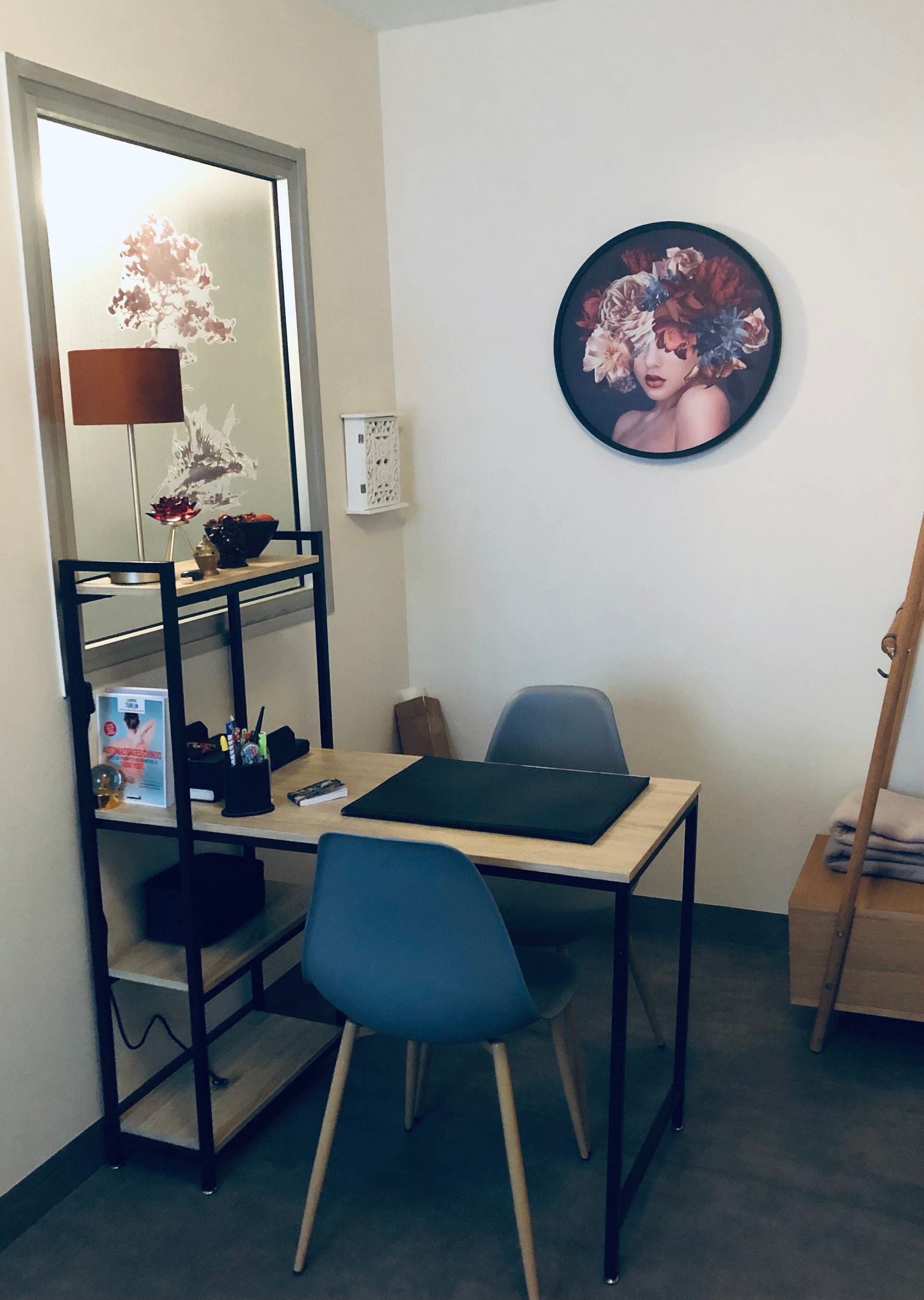 Bureau dans un bureau avec une chaise bleue, une œuvre d'art ronde représentant le visage d'une femme, une étagère et une fenêtre.