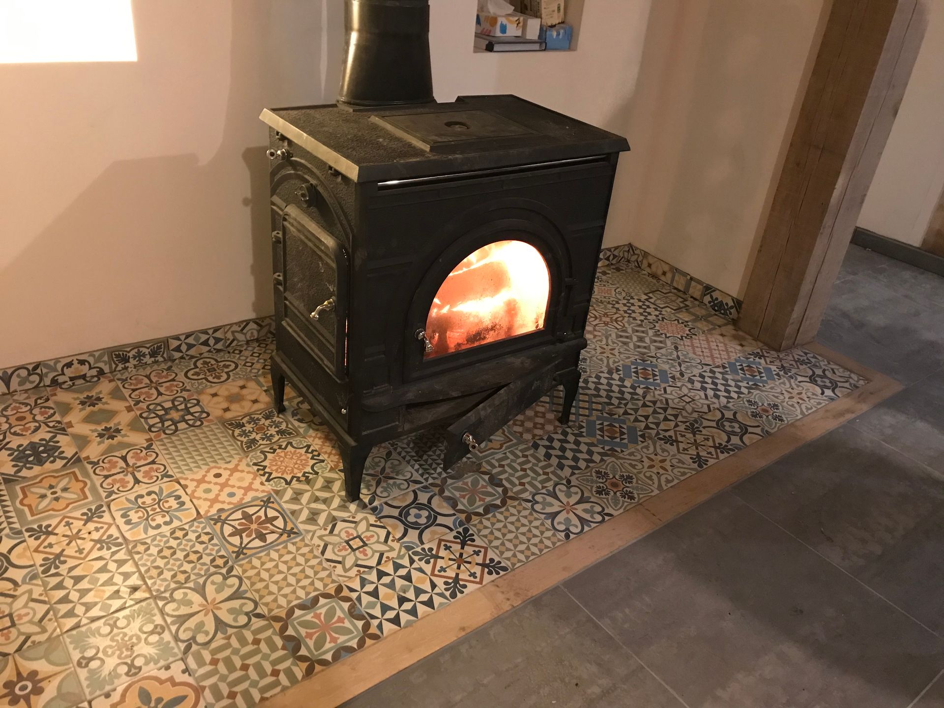 Un poêle à bois noir avec un feu allumé est posé sur un sol carrelé à motifs dans une pièce.