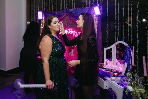 En una fiesta, dos mujeres se miran fijamente bajo luces violetas; una de ellas sostiene un micrófono.