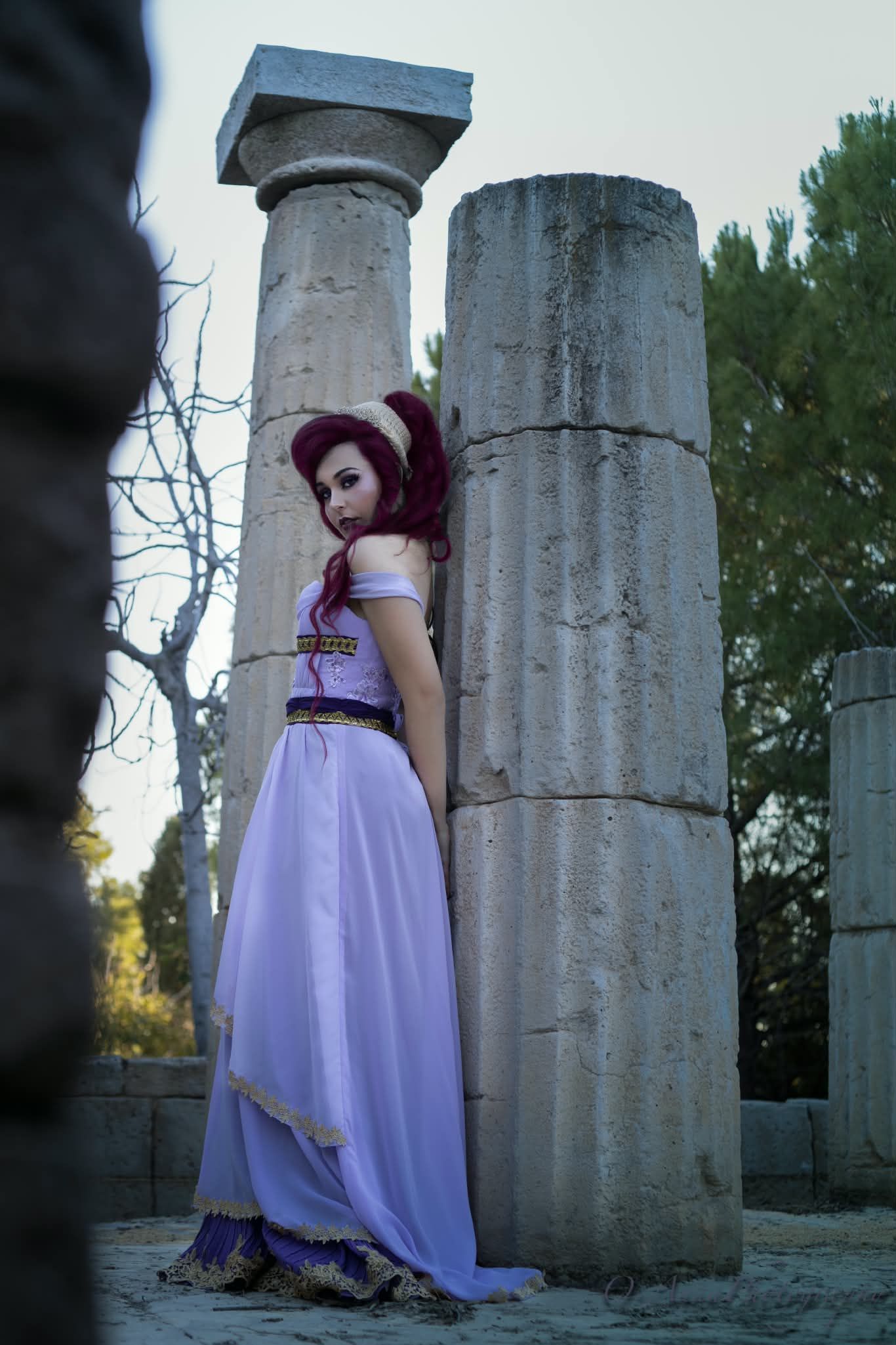Una persona con un vestido color lavanda está de pie entre columnas de piedra al aire libre al anochecer.