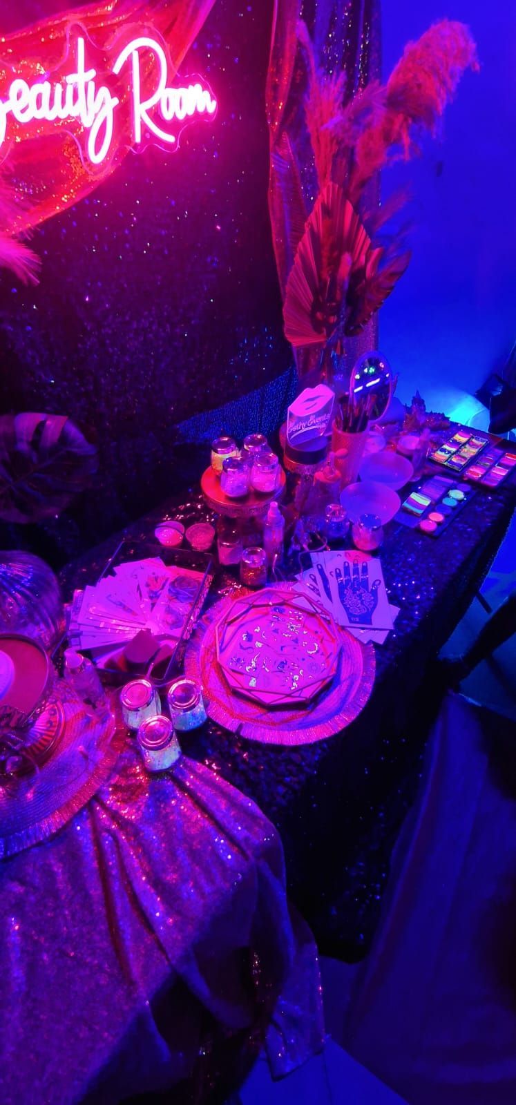 Mesa de fiesta iluminada con luces de neón de color rosa y morado, flores, postres, bebidas y un letrero luminoso que dice