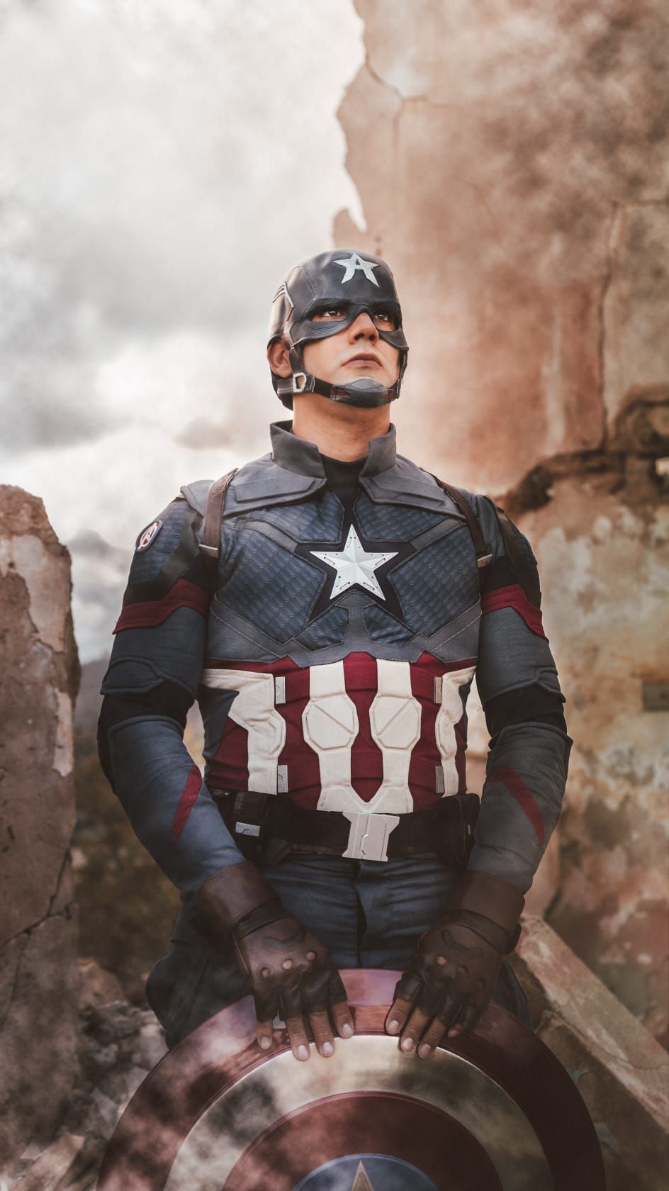 El Capitán América, ataviado con su armadura de combate, mira hacia arriba entre el humo y los escombros, sosteniendo su escudo.