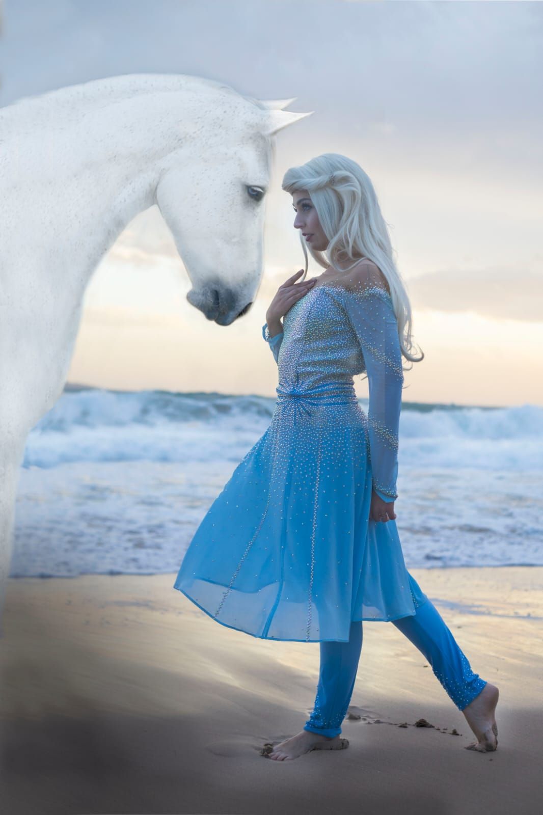 Una mujer con un vestido azul mira a un caballo blanco en una playa al atardecer.