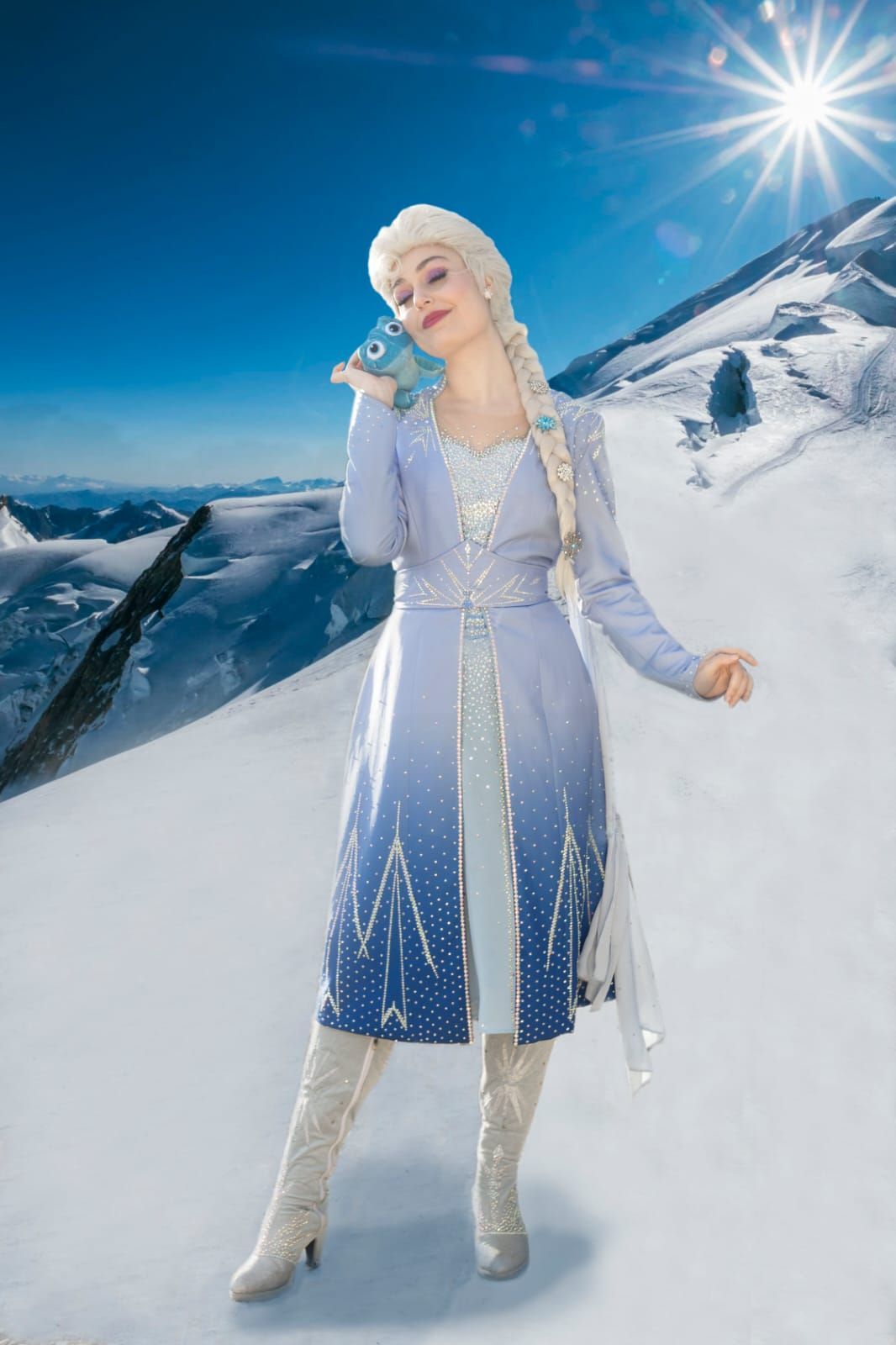 Mujer con un vestido de invierno azul pálido sosteniendo un micrófono en montañas nevadas bajo un sol radiante.