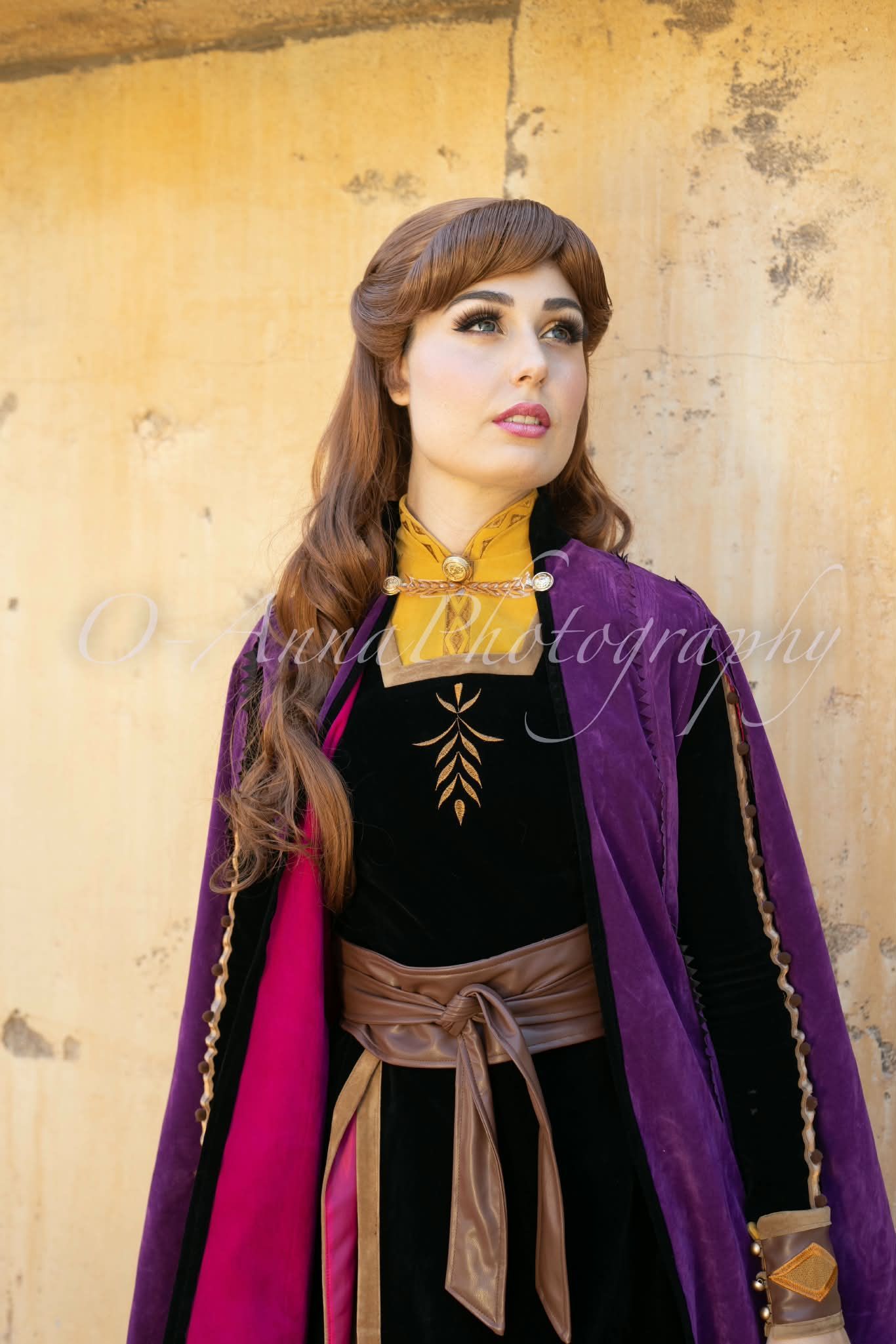 Persona con abrigo morado y ropa negra de pie contra una pared amarilla, mirando hacia un lado.