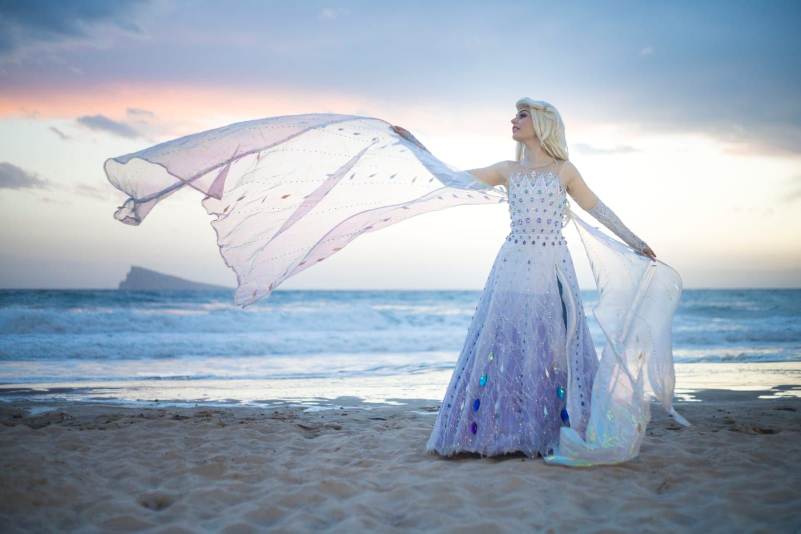 Mujer con un vaporoso vestido color lavanda sosteniendo una tela en una playa al atardecer.