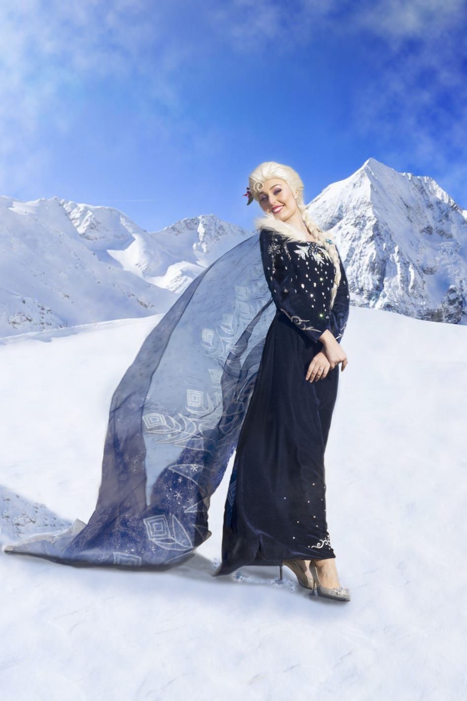 Mujer con un vestido negro y una capa azul ondeante de pie sobre montañas nevadas bajo un cielo azul brillante