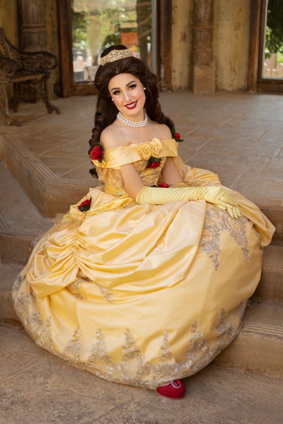 Mujer con un vestido de gala amarillo sentada en un patio de piedra, luciendo joyas de perlas y zapatos rojos.