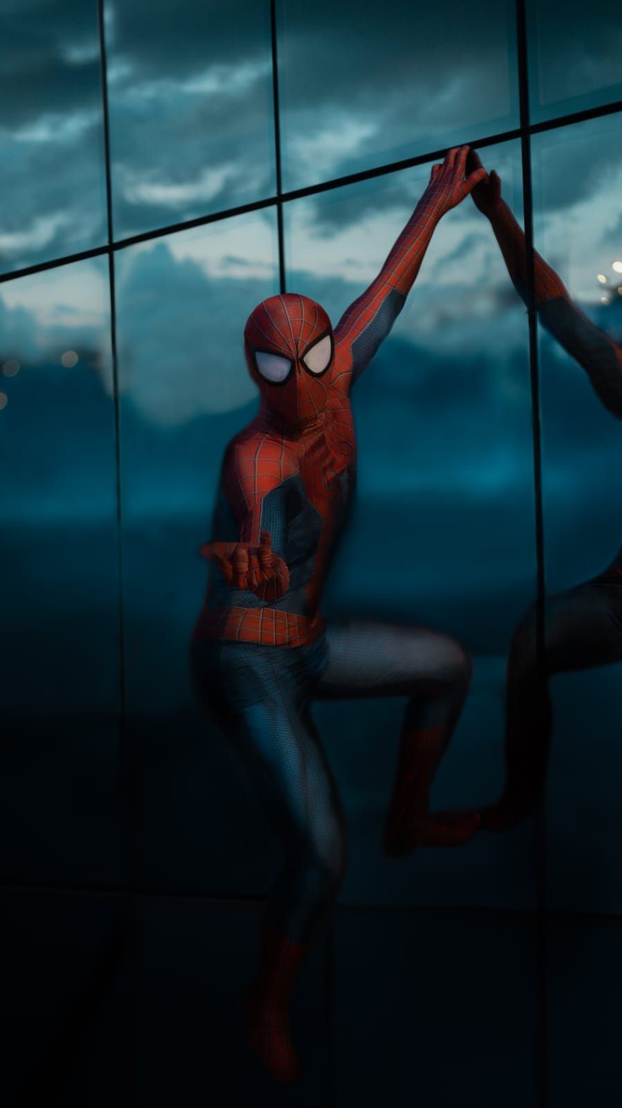 Spider-Man posando sobre la pared reflectante de un edificio, con un cielo azul nublado de fondo.