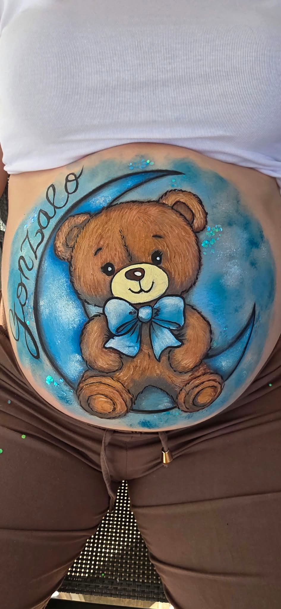 Pintura redonda de un osito de peluche con una cinta azul sobre el vientre de una mujer embarazada, con remolinos azules.