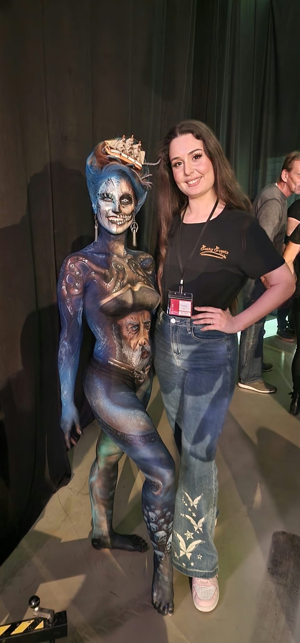Dos personas posando juntas en un espacio para eventos con poca luz, una con el cuerpo pintado de azul metalizado y la otra con camisa negra y vaqueros.