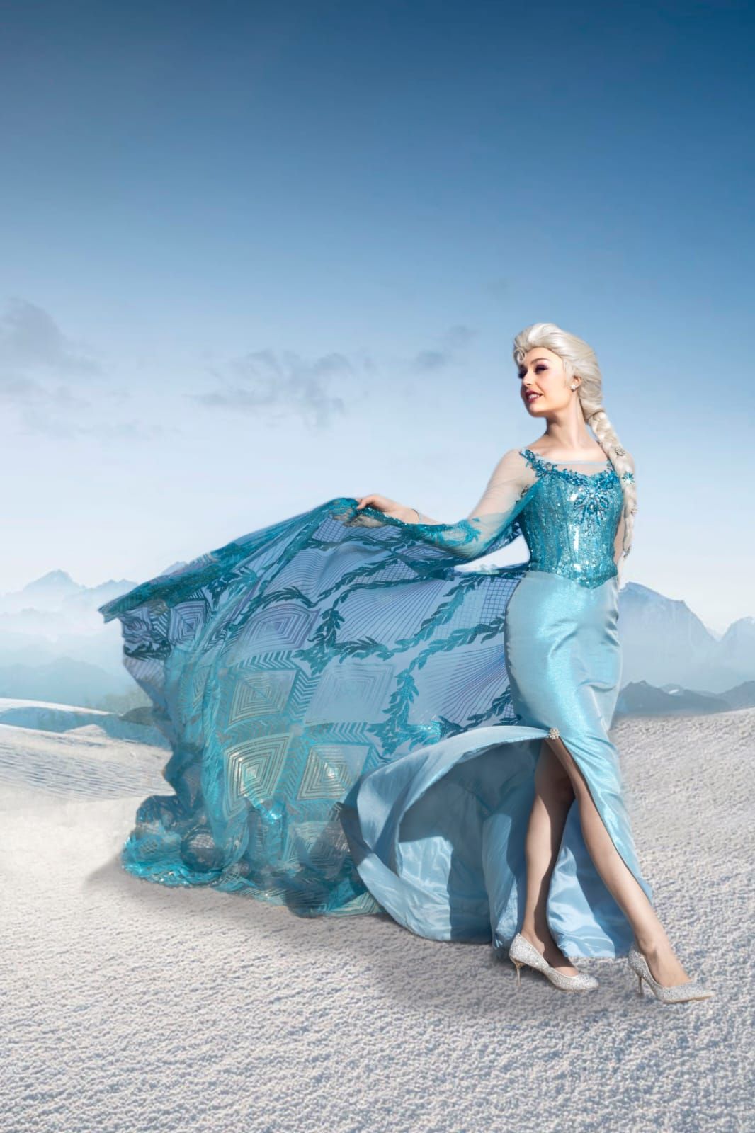 Mujer con un vaporoso vestido azul de pie en un paisaje nevado con montañas en la distancia.