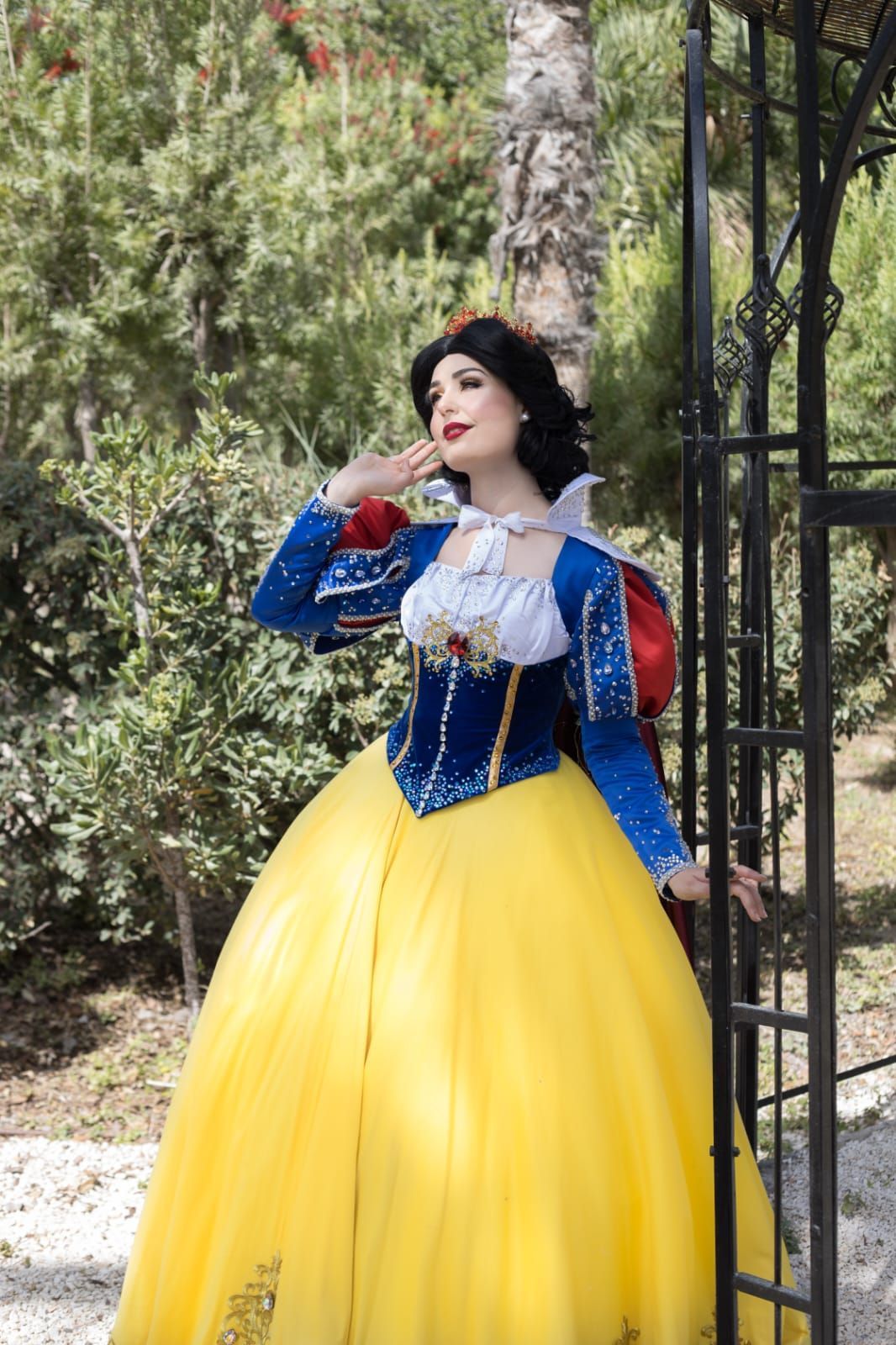 Mujer disfrazada de Blancanieves posando al aire libre junto a una verja metálica y árboles.