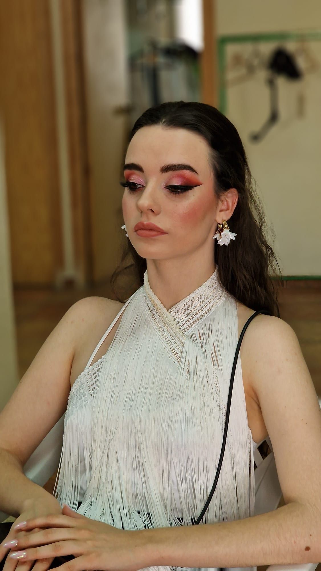 Mujer con un vestido blanco con cuentas y maquillaje rosa en los ojos, sentada en el interior y mirando hacia abajo.