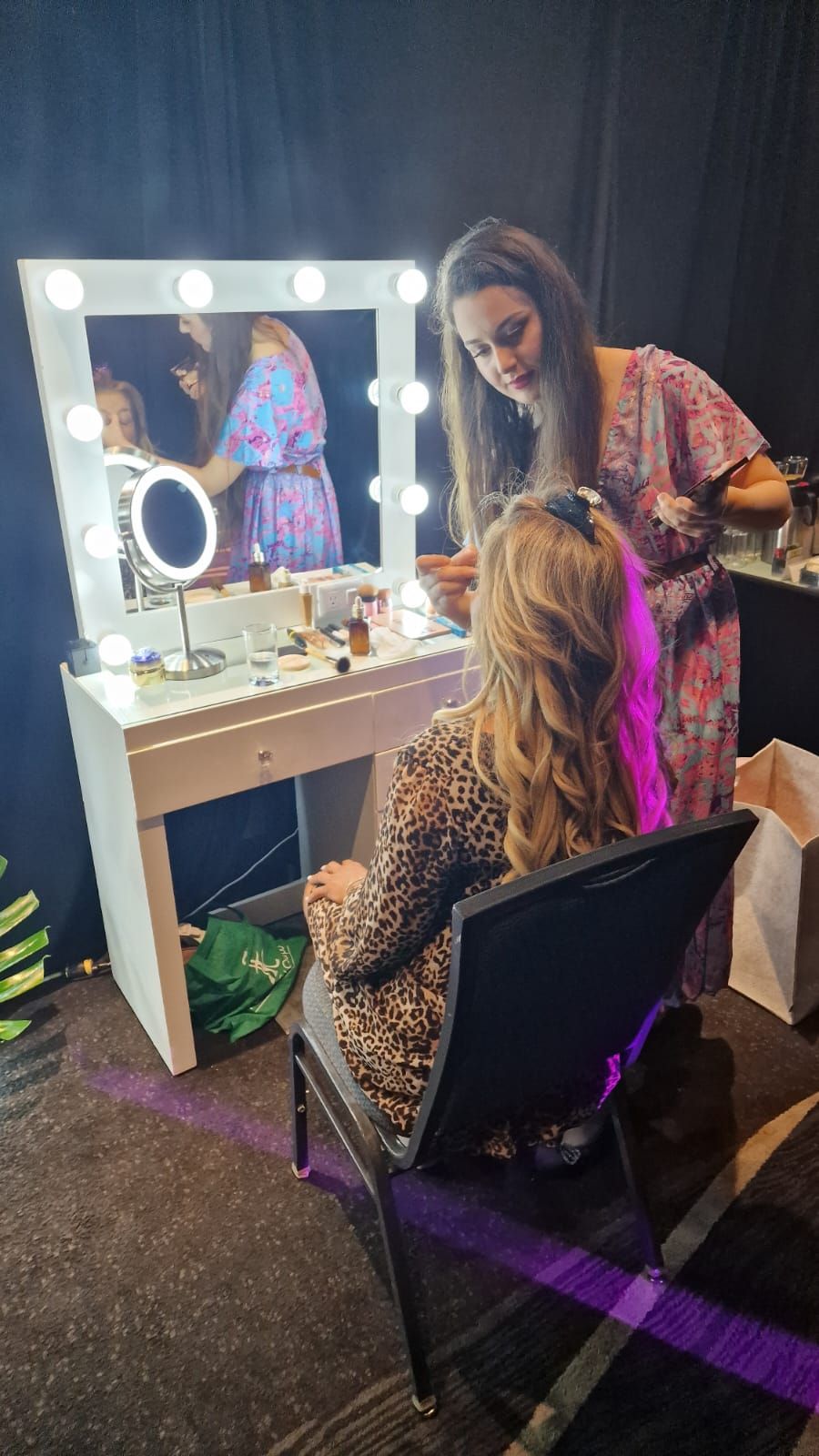 Una mujer peinando a otra frente a un espejo de tocador con luces en un vestidor.