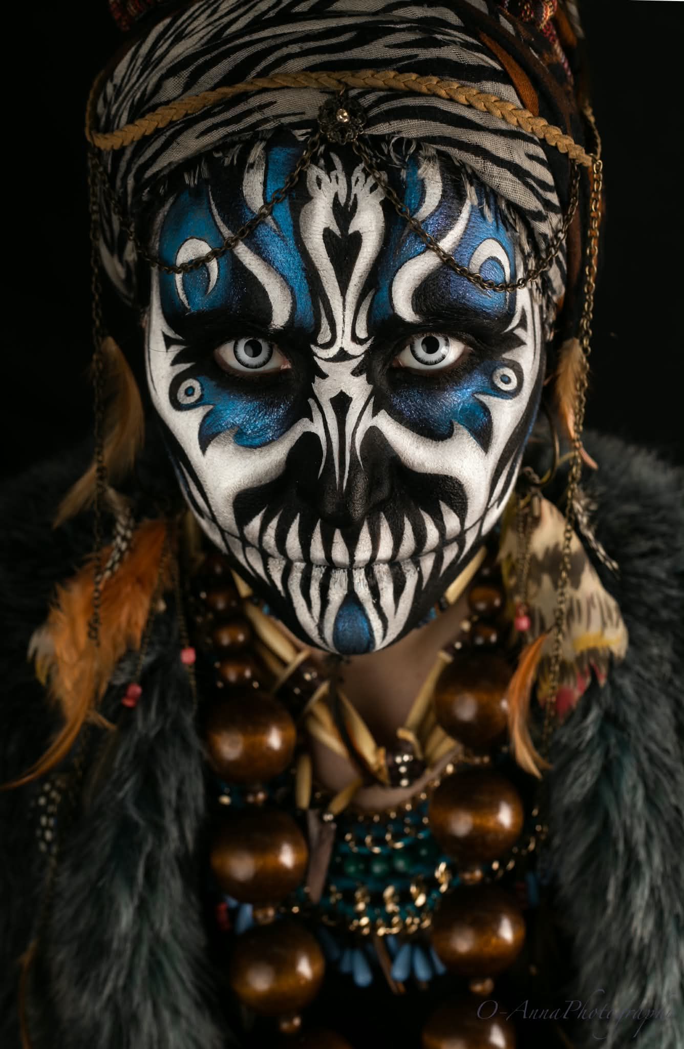 Rostro pintado con maquillaje de calavera azul y blanco, con turbante y collar de cuentas sobre un fondo oscuro.
