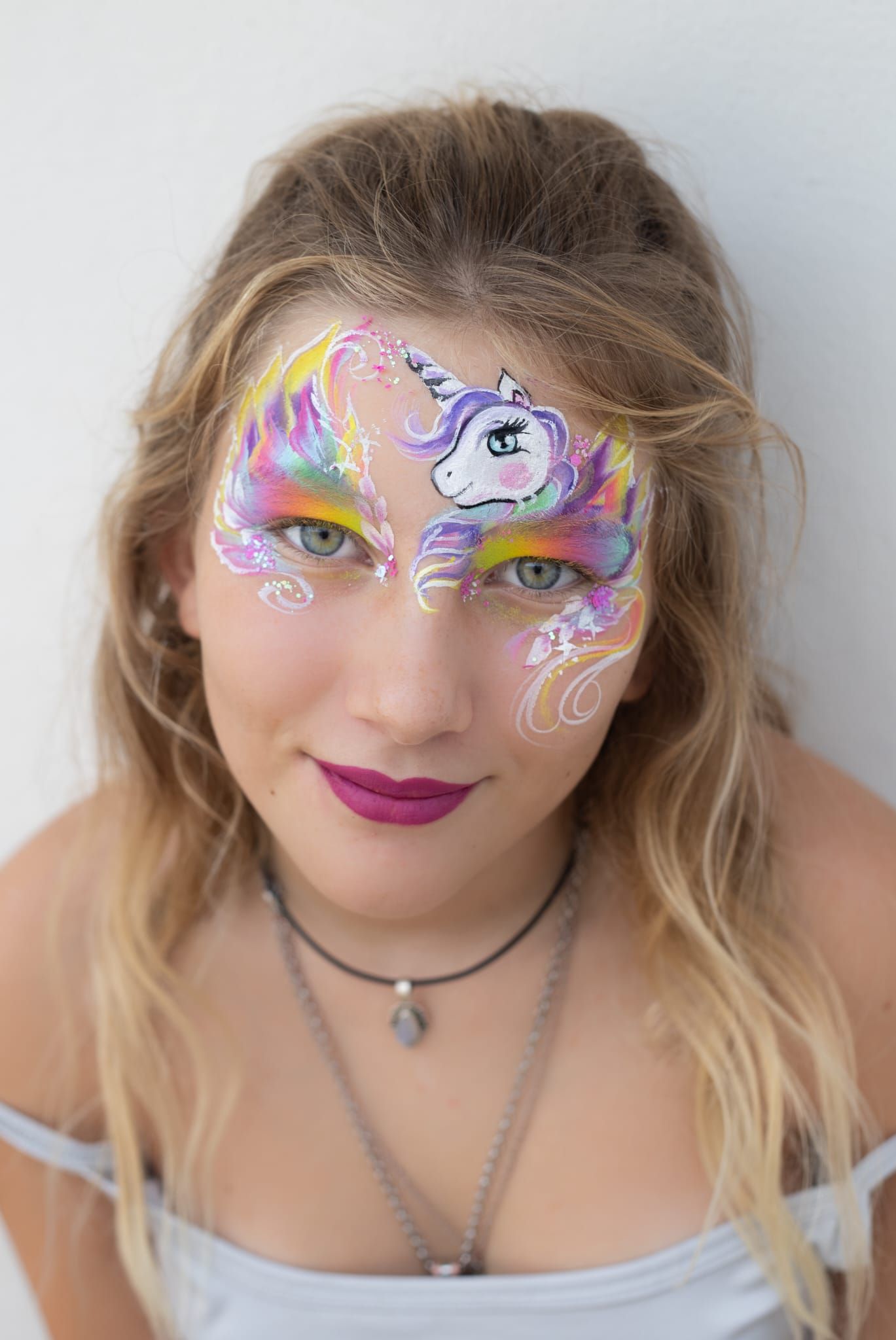 Mujer con la cara pintada de colores y maquillaje de mariposa, vestida con una blusa blanca y un collar, mirando a la cámara.