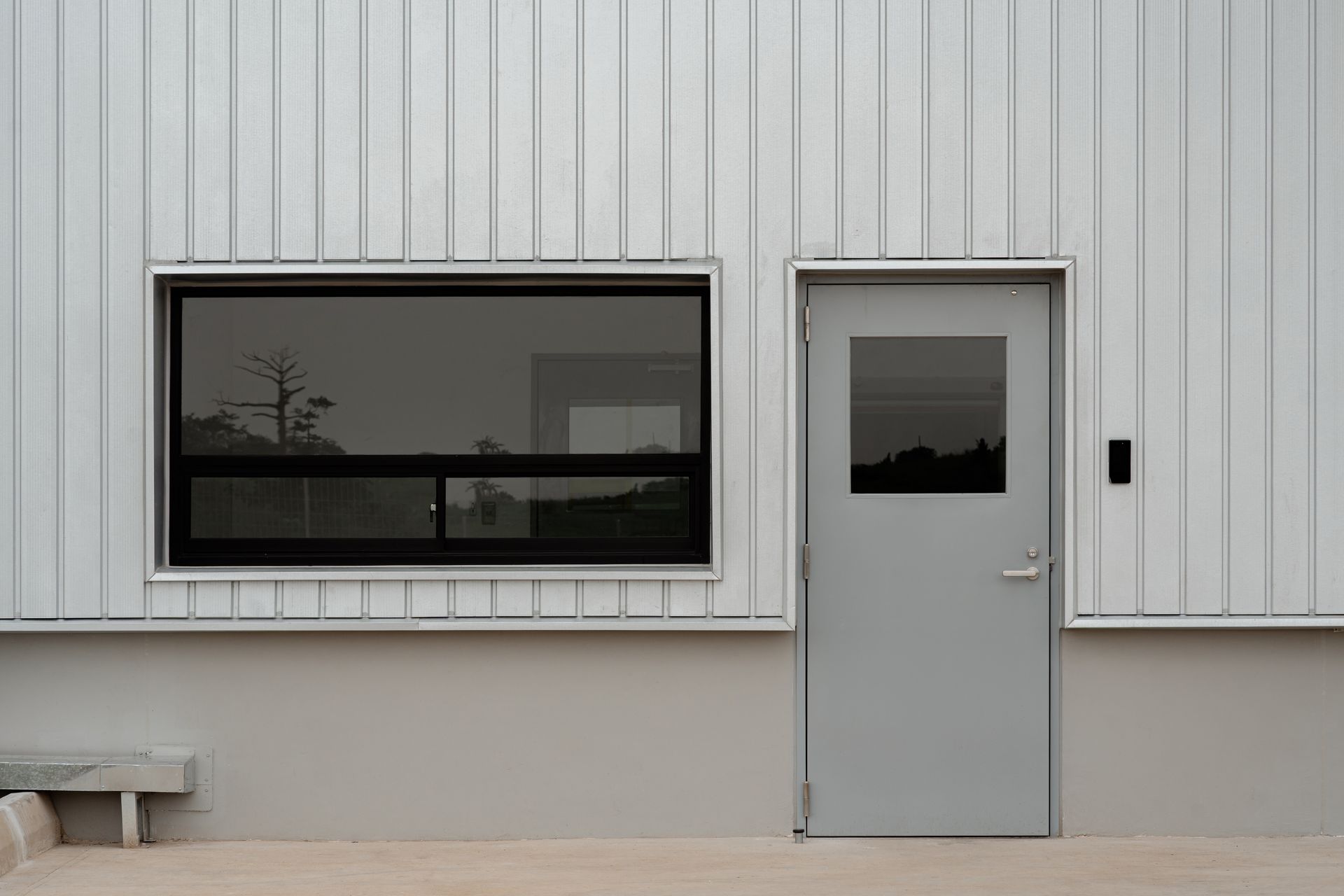 Façade grise du bâtiment avec une grande fenêtre sombre et une porte grise sur un mur à bardage blanc