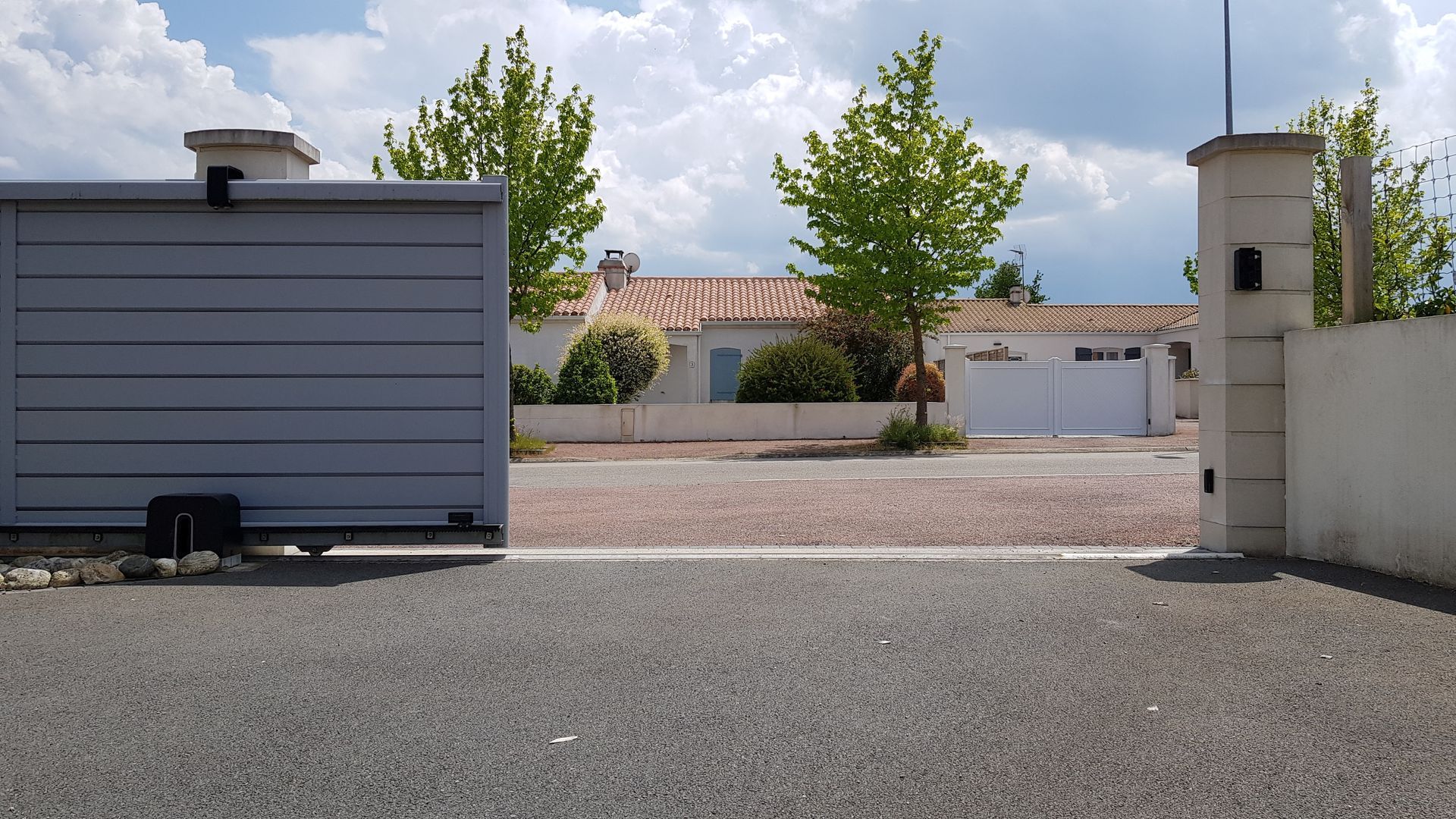 Allée de gravier menant à une maison moderne aux murs gris, portail blanc et arbres sous un ciel nuageux