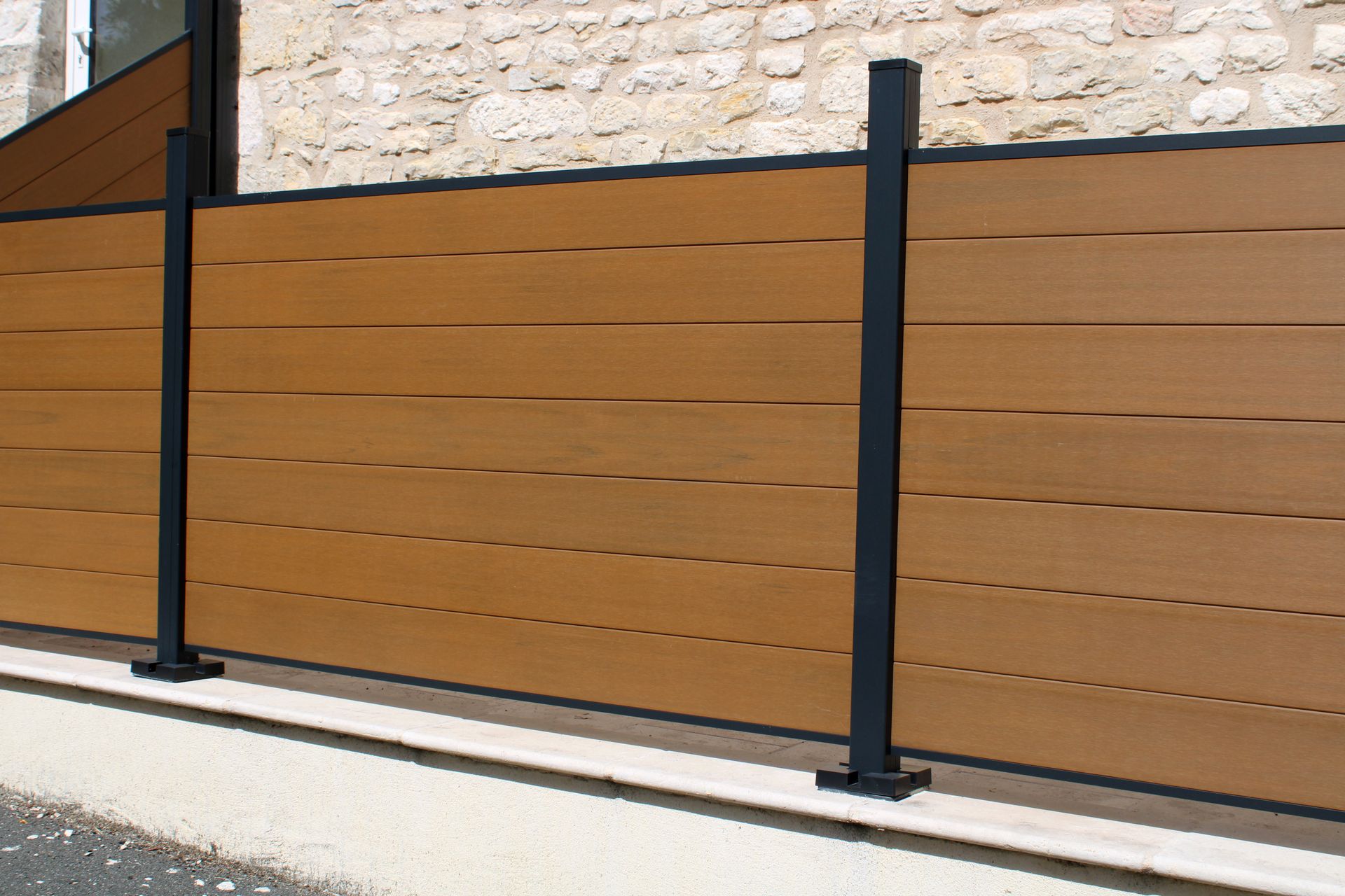 Clôture moderne à lattes brunes avec poteaux noirs, le long d'un mur en pierre