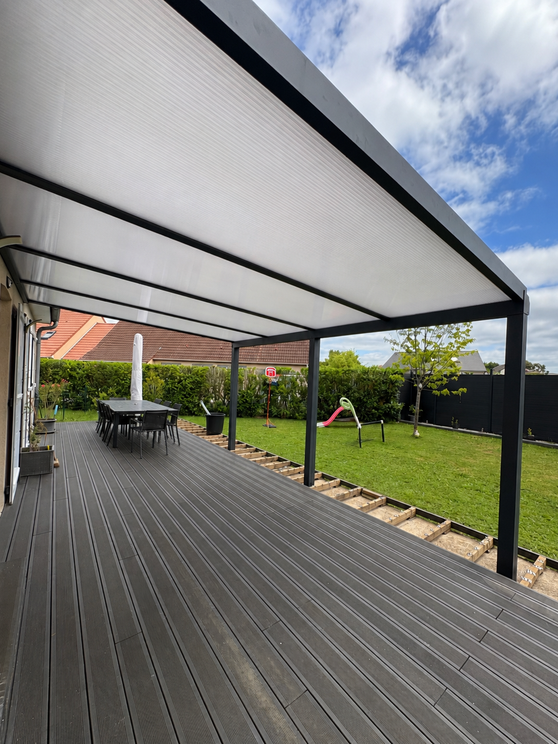 Terrasse en bois couverte jouxtant une cour gazonnée, avec des poteaux noirs et une table sous une pergola