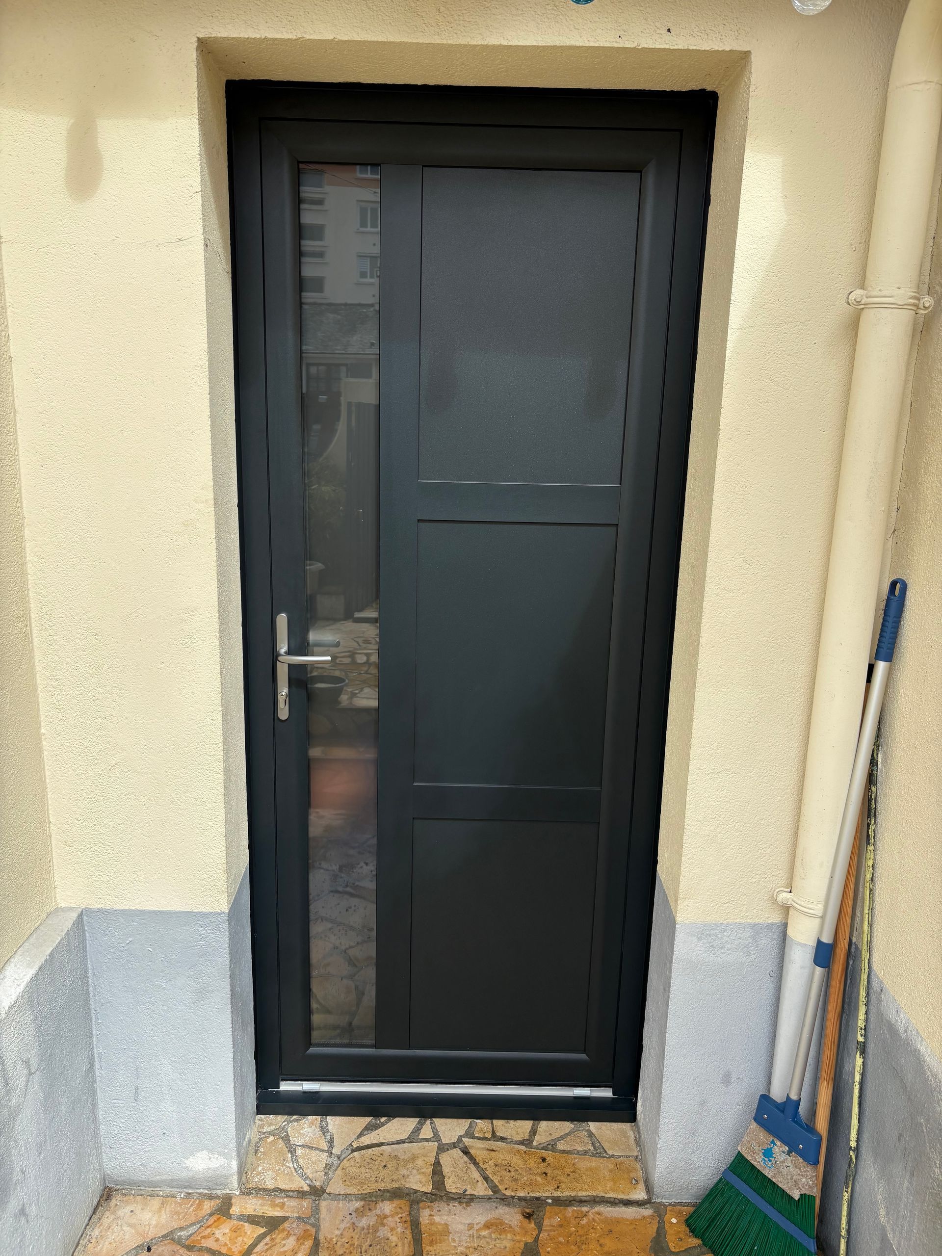 Porte en métal noir sur un mur beige, avec un balai appuyé à côté