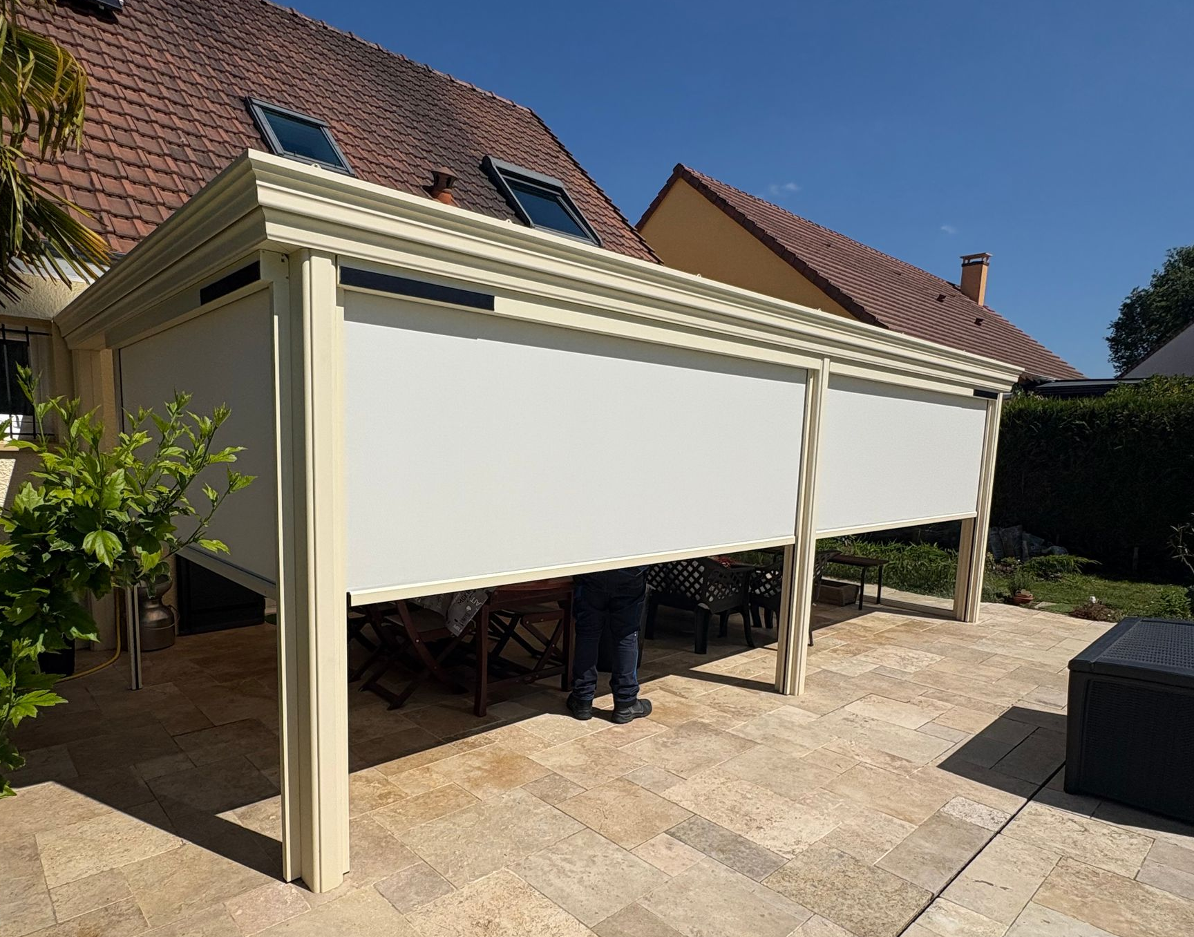 Store beige rétractable couvrant une pergola, partiellement abaissé, avec une personne debout en dessous, à côté d'une maison
