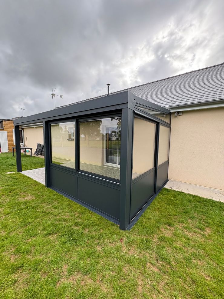 Véranda en aluminium noir attenante à une maison beige, avec un jardin gazonné et un ciel nuageux