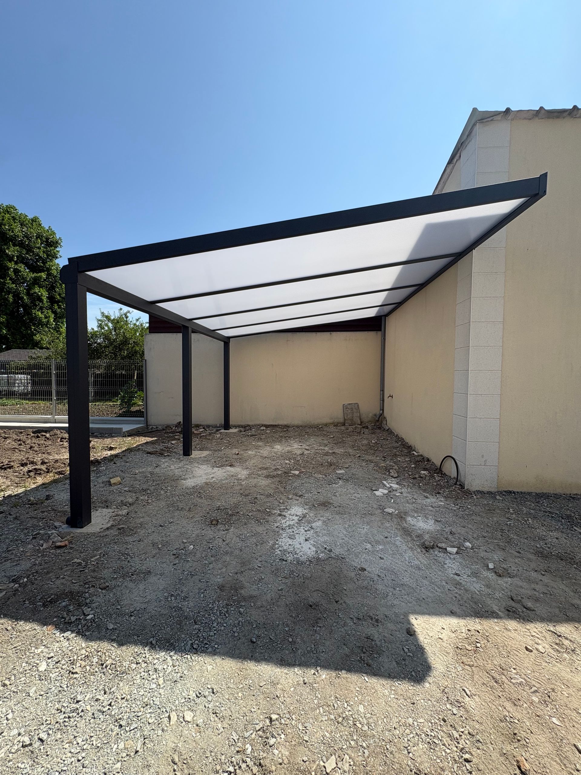 Abri de voiture, à côté d'un bâtiment beige, avec des supports en métal noir et un toit translucide