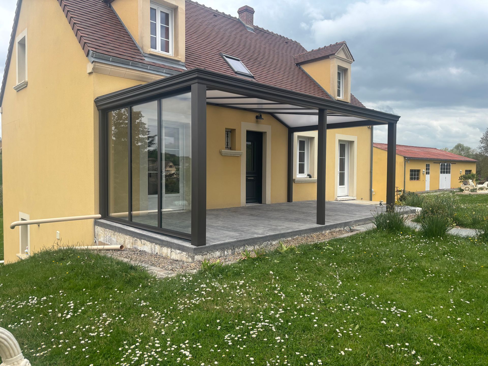 Maison jaune avec véranda, à côté d'une pelouse verte parsemée de fleurs blanches.