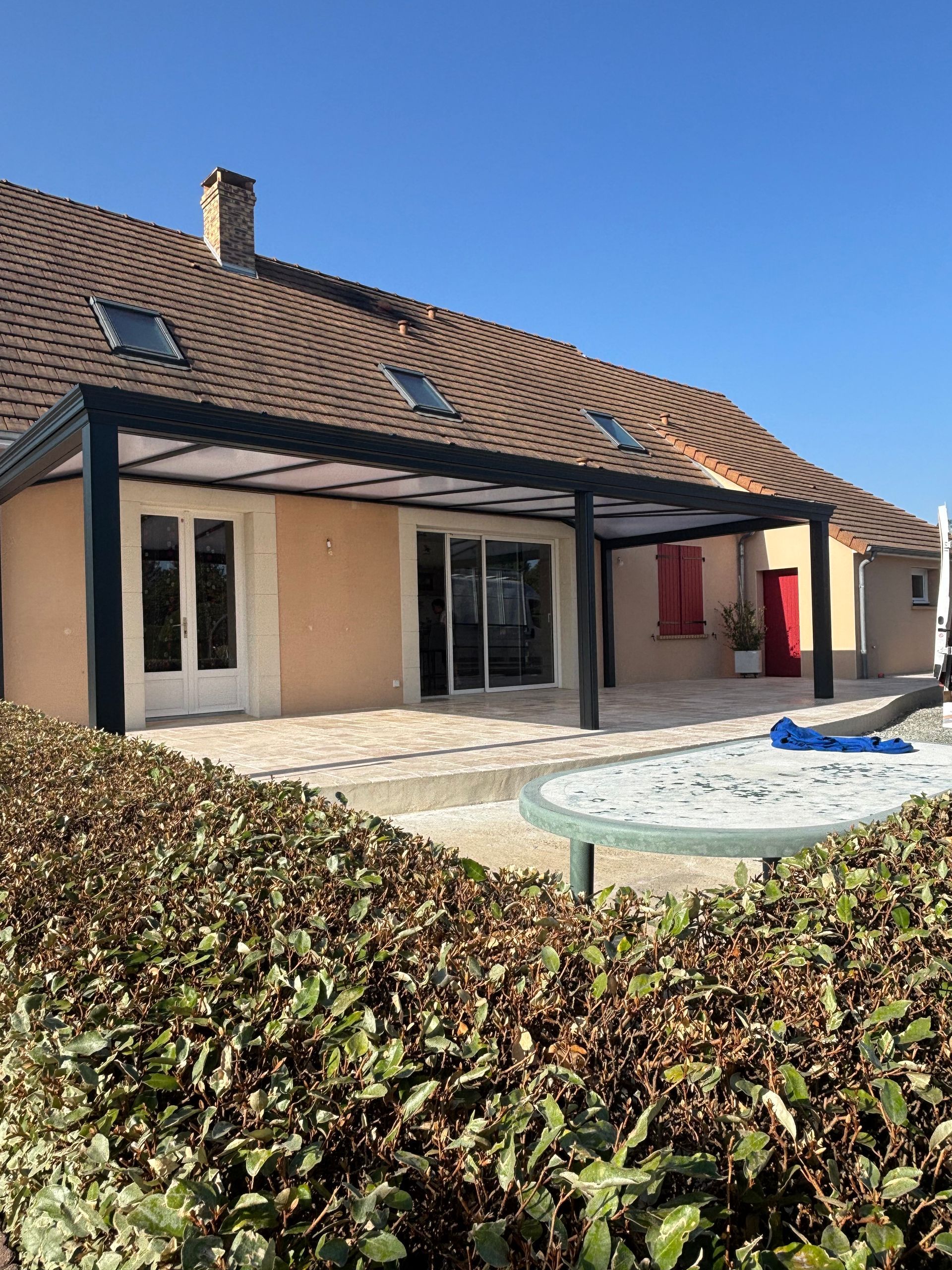 Patio ensoleillé avec une maison beige, toit de tuiles, grandes fenêtres et une haie au premier plan