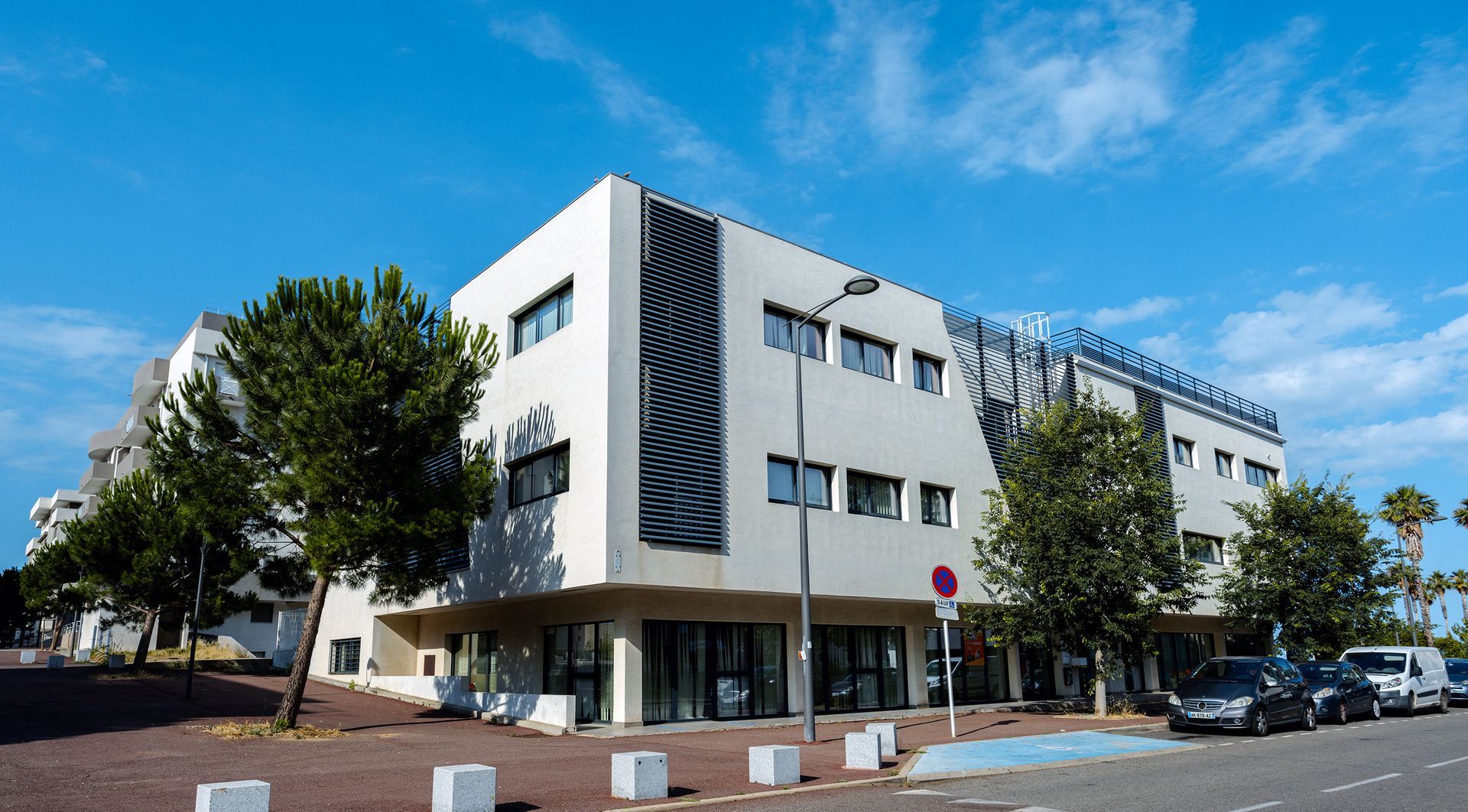 Bâtiment moderne et blanc avec des accents sombres, de petits arbres et des voitures garées sous un ciel bleu.