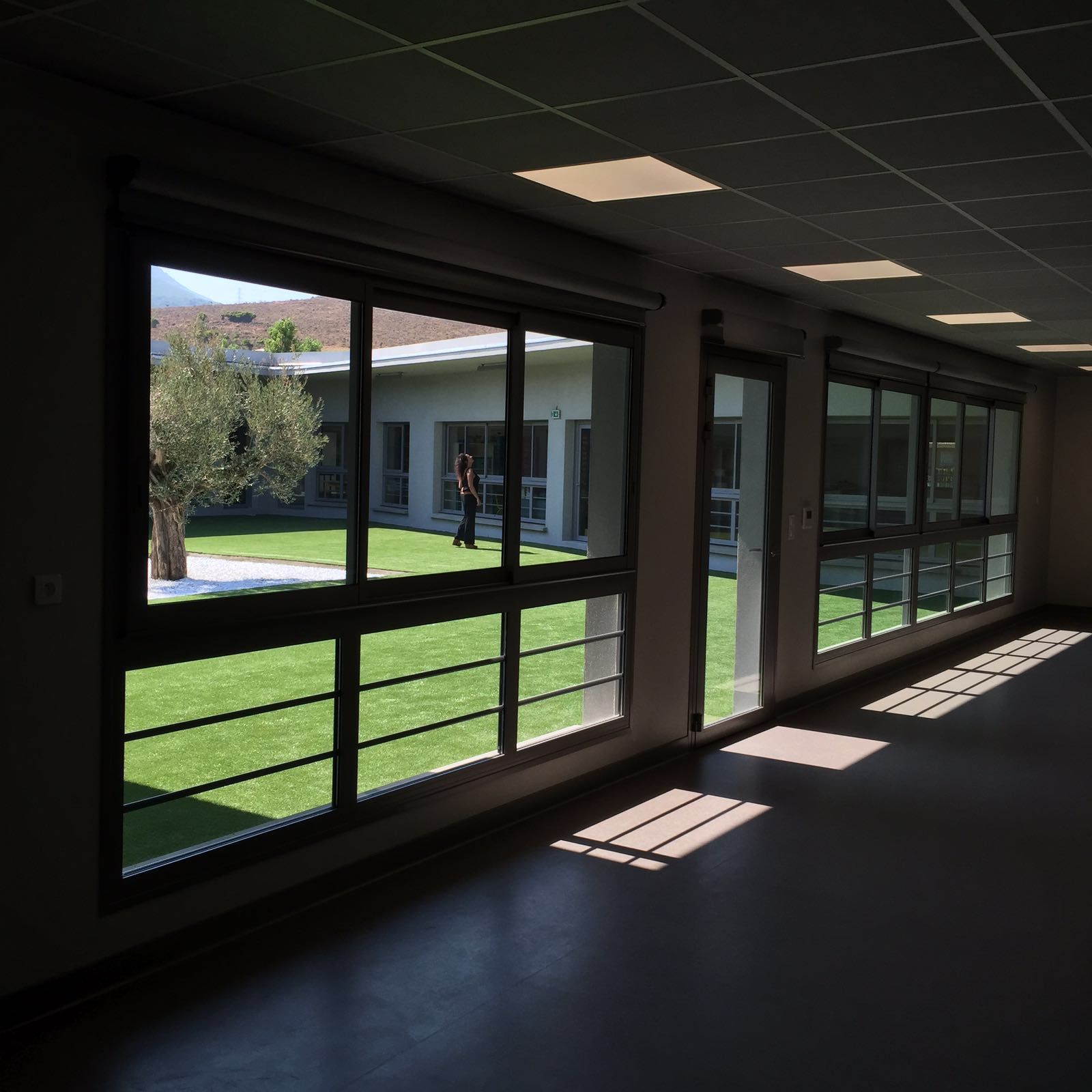 Pièce intérieure avec de grandes fenêtres donnant sur une cour verdoyante et un bâtiment.