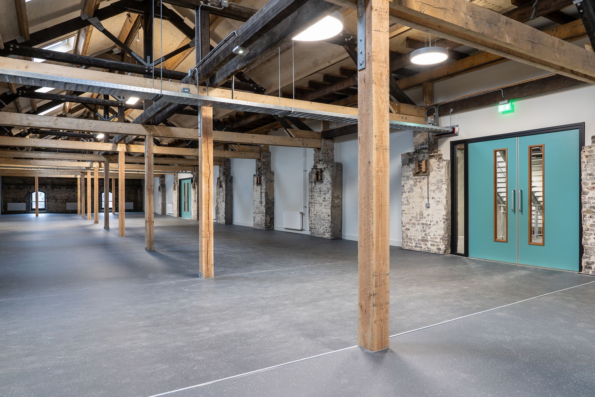 Pièce vide de style industriel avec poutres en bois, murs en briques et sol gris ; portes doubles bleu sarcelle à droite.