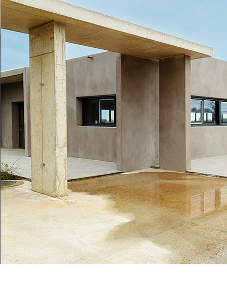 Extérieur d'un bâtiment moderne avec des murs beiges, des fenêtres sombres et un surplomb en béton se reflétant dans une flaque d'eau.